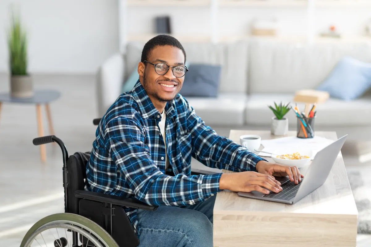 Un hombre sonriente con gafas está sentado en su silla de ruedas frente a un escritorio, con las manos sobre un ordenador portátil, mientras realiza un examen diseñado para la accesibilidad digital.