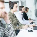 オンラインでのリアルタイム監視の概念を示すため、ヘッドセットを装着し、一列に並んでコンピューター画面を見つめるヘルプデスクオペレーター数名の横顔