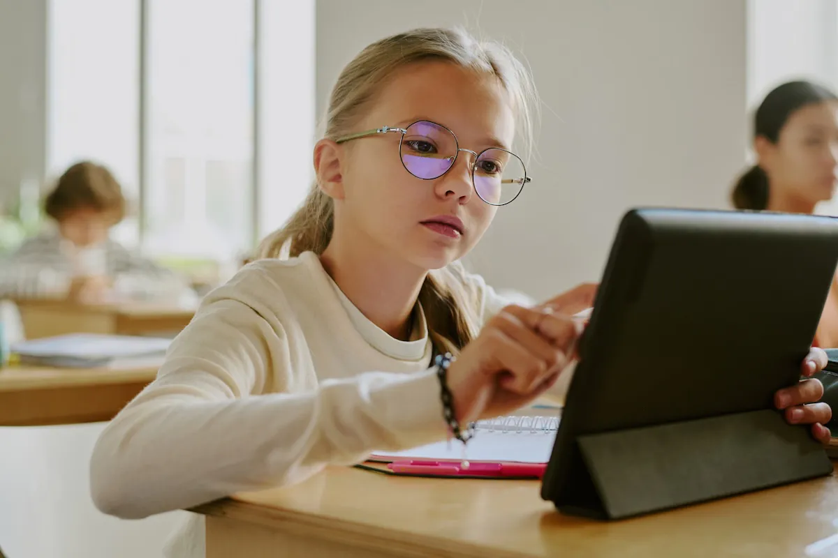 Una ragazzina con gli occhiali seduta a un banco di legno mentre usa un tablet in un'aula circondata dai compagni di classe